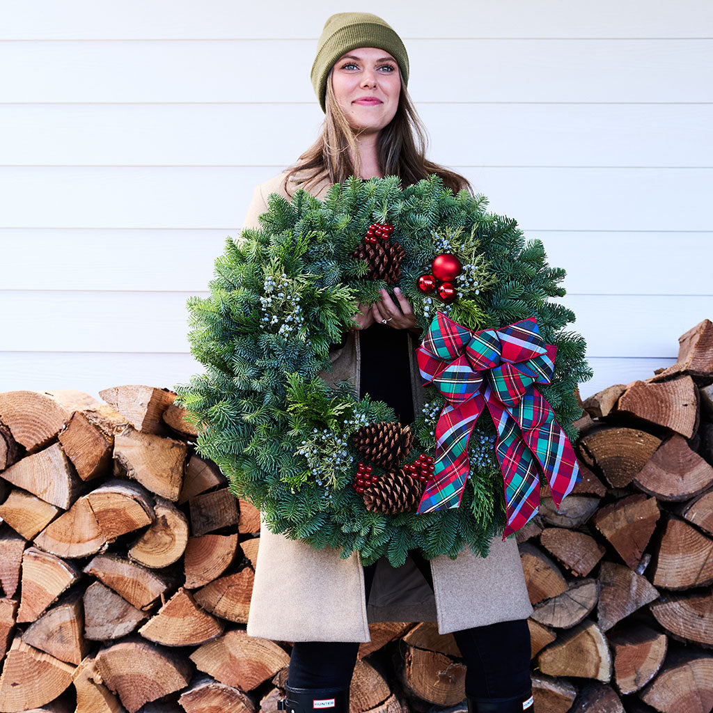 A woman wearing an olive green beanie and a beige coat holds a large green Christmas wreath decorated with pinecones, red berries, and a red, green, and blue plaid bow. She is standing in front of a white wood-paneled wall and a stacked firewood pile.