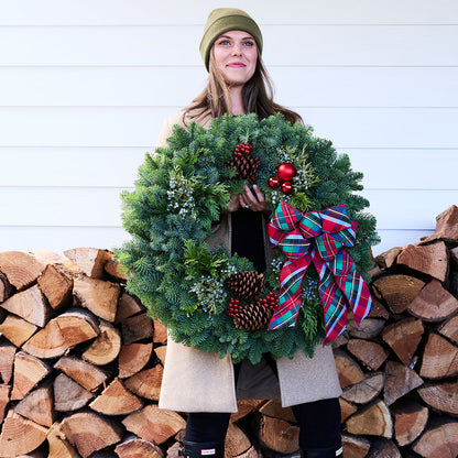 A woman wearing an olive green beanie and a beige coat holds a large green Christmas wreath decorated with pinecones, red berries, and a red, green, and blue plaid bow. She is standing in front of a white wood-paneled wall and a stacked firewood pile.