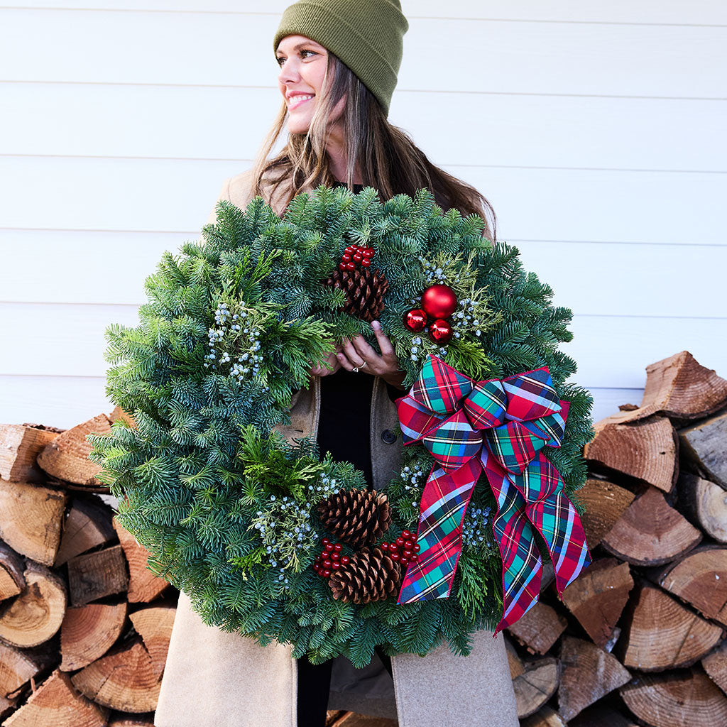 A woman wearing a green beanie and beige coat holds a large green Christmas wreath decorated with pinecones, red berries, and a red plaid bow, in front of a white paneled wall and stacked firewood.