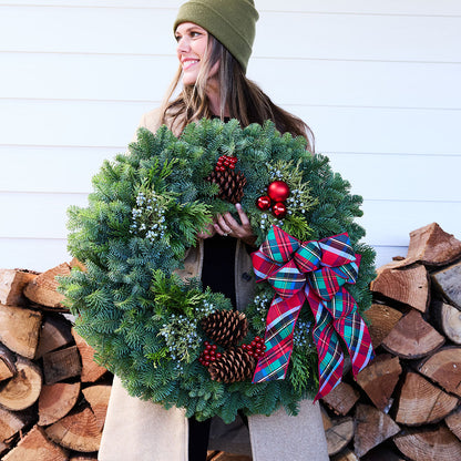 A woman wearing a green beanie and beige coat holds a large green Christmas wreath decorated with pinecones, red berries, and a red plaid bow, in front of a white paneled wall and stacked firewood.