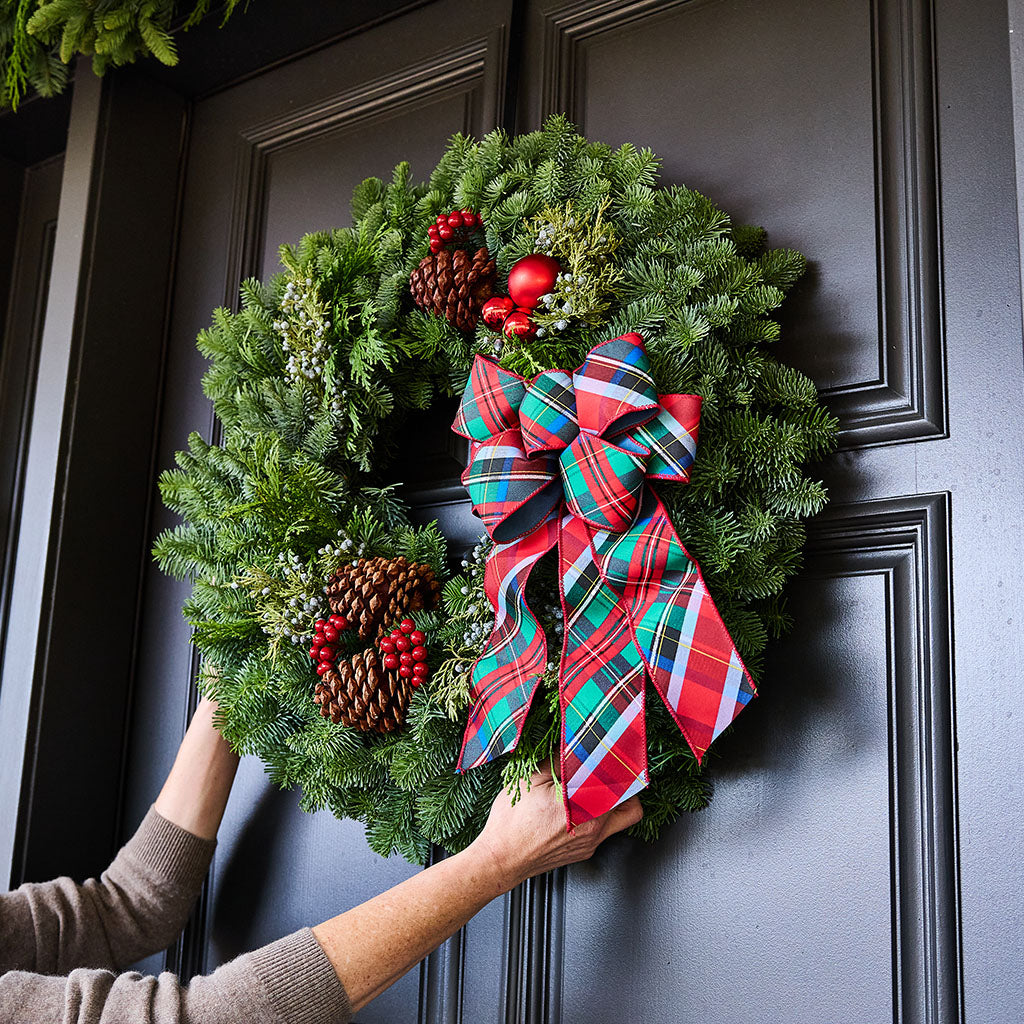 A green evergreen wreath with pinecones, red berries, and a large red plaid bow being held against a dark paneled door by a person's hands and arms.