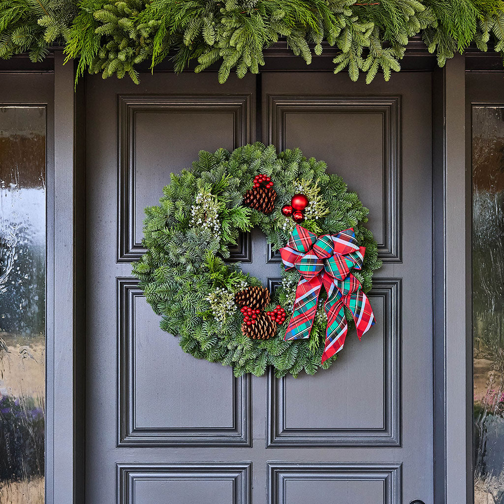 A green pine wreath with red berries, pinecones, and a red, green, and blue plaid bow hanging on a dark gray front door, with a green garland above.