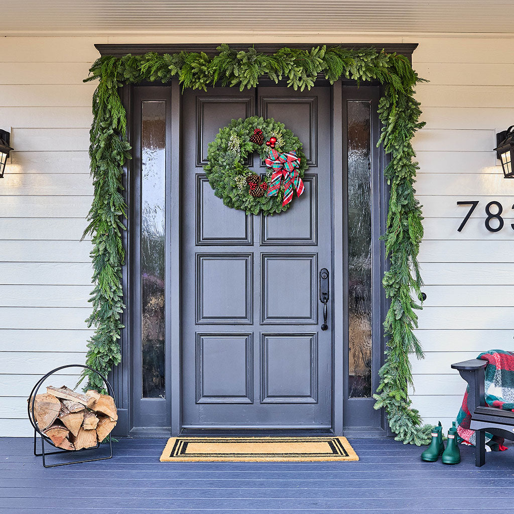 A front door decorated for Christmas with a green garland arch, a wreath with red and green ribbon, a doormat, a log burner, and a bench with a plaid throw on a white siding porch.