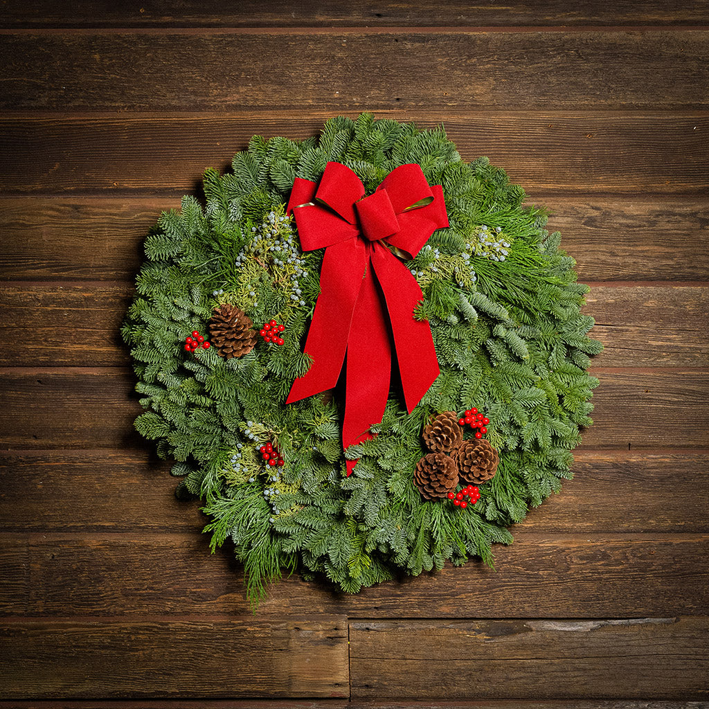 An evergreen wreath of noble fir and juniper with natural pinecones, faux red berries, and a red velvet bow