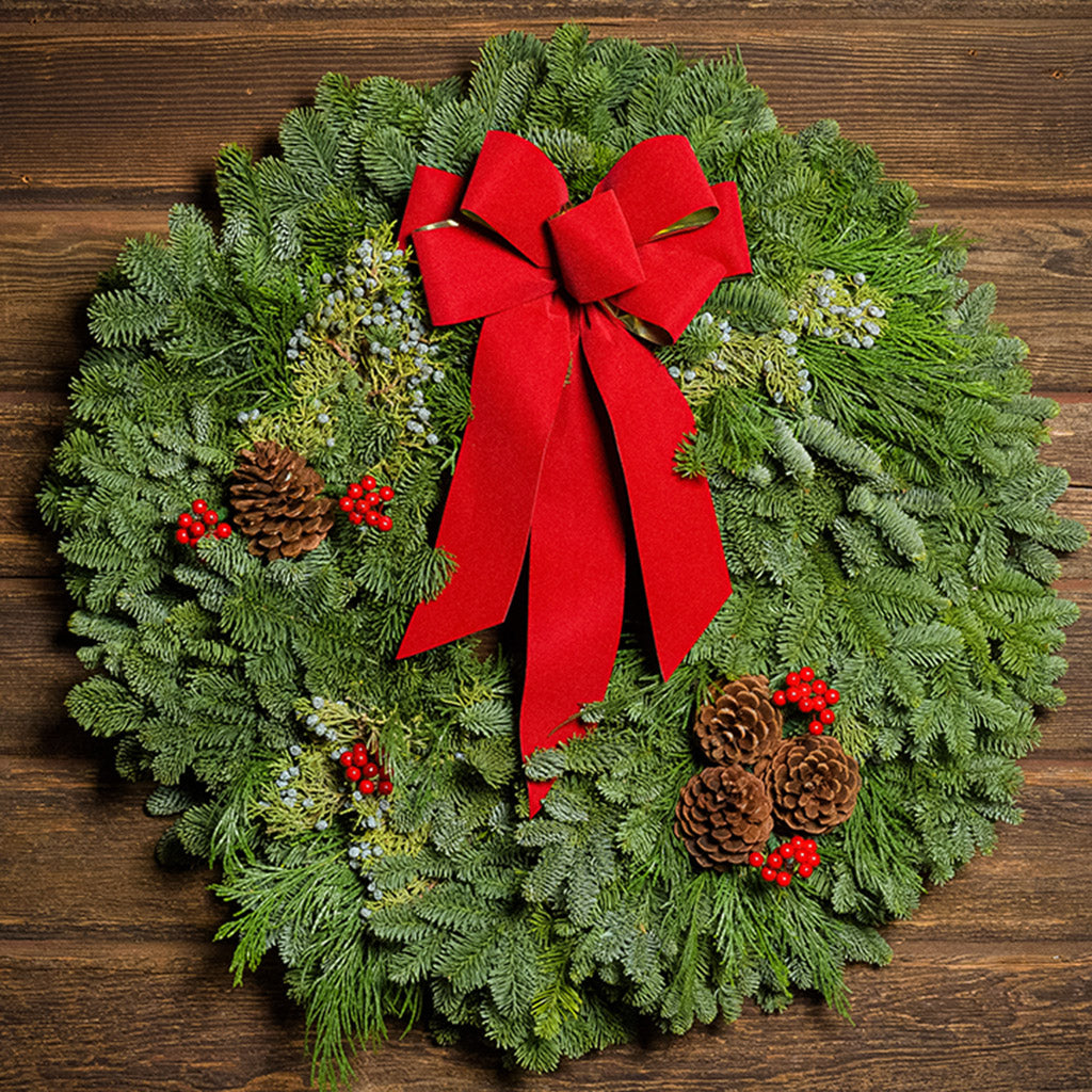 An evergreen wreath of noble fir and juniper with natural pinecones, faux red berries, and a red velvet bow