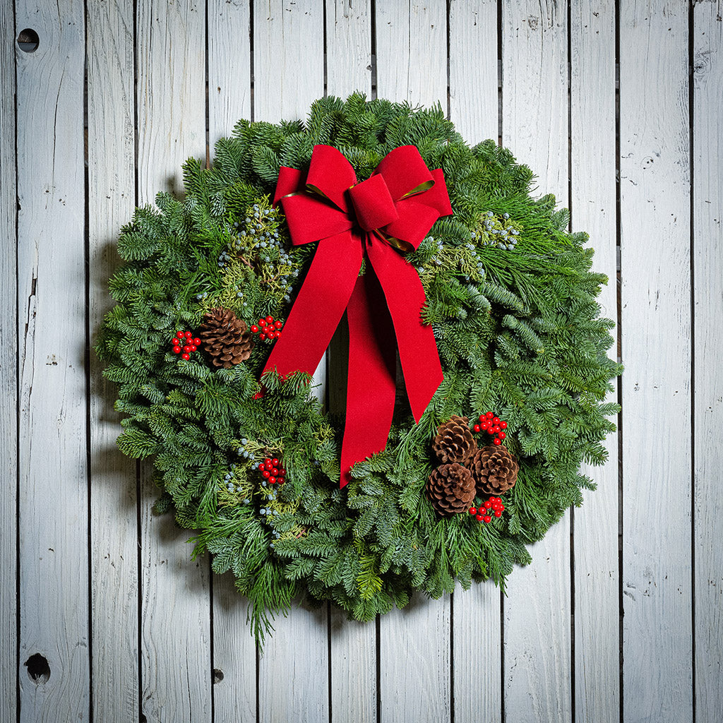 An evergreen wreath of noble fir and juniper with natural pinecones, faux red berries, and a red velvet bow