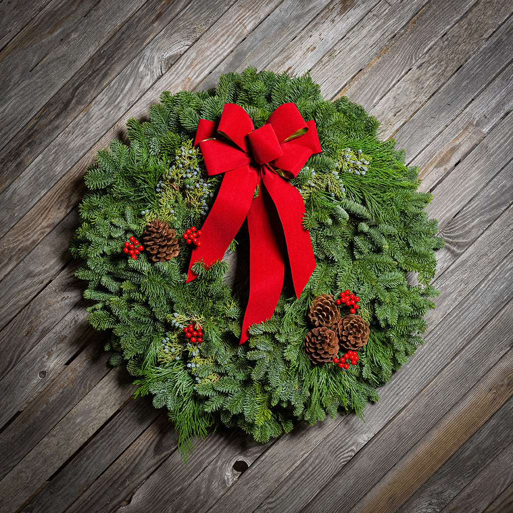 An evergreen wreath of noble fir and juniper with natural pinecones, faux red berries, and a red velvet bow