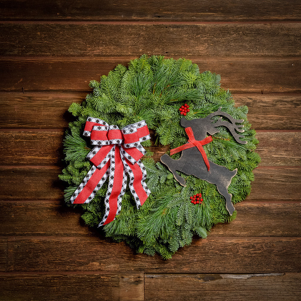 A wreath made of noble fir and pine with red berries, a black leaping deer cutout, and a red with black and white edged plaid bow hanging on a dark wooden background.