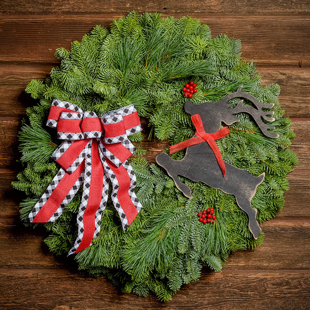 A wreath made of noble fir and pine with red berries, a black leaping deer cutout, and a red with black and white edged plaid bow hanging on a dark wooden background.