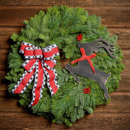 A wreath made of noble fir and pine with red berries, a black leaping deer cutout, and a red with black and white edged plaid bow hanging on a dark wooden background.