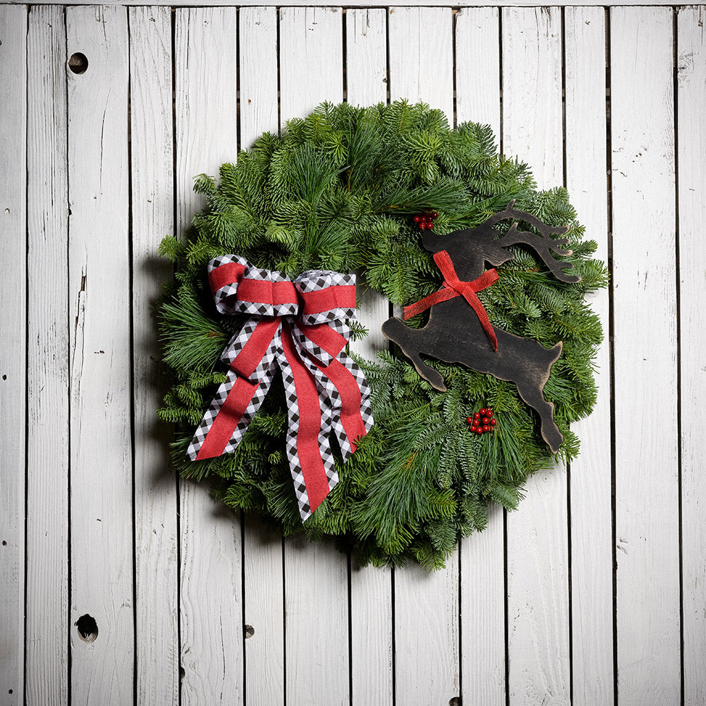 A wreath made of noble fir and pine with red berries, a black leaping deer cutout, and a red with black and white edged plaid bow hanging on a dark wooden background.