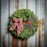 A wreath made of noble fir and pine with red berries, a black leaping deer cutout, and a red with black and white edged plaid bow hanging on a dark wooden background.