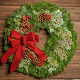 A close-up of a wreath made of silver fir and juniper that is decorated with faux red berries and small country apples, ponderosa pinecones, and a red velveteen with gold back bow hung on dark wood background.