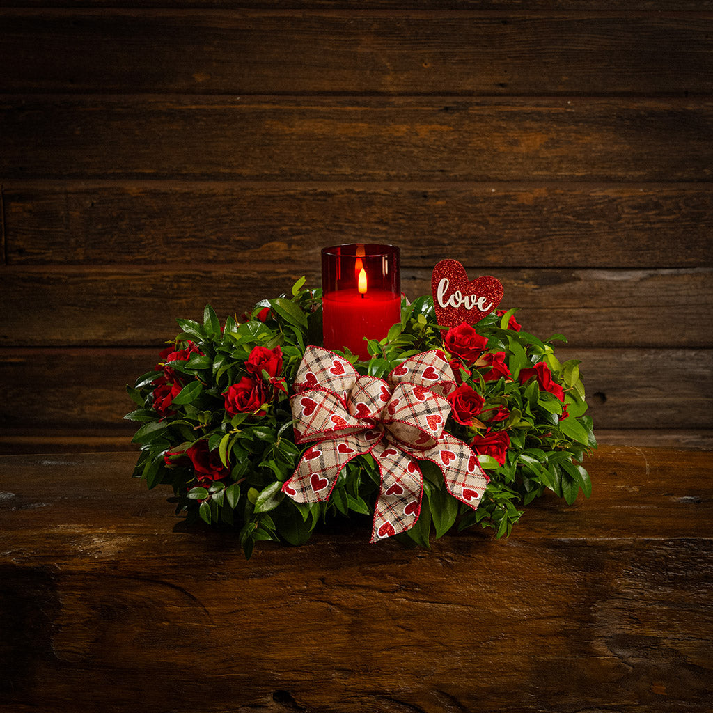 Fresh salal and green huckleberry with faux mini red roses, a red heart “love” sign, a red heart bow, and a red LED candle.