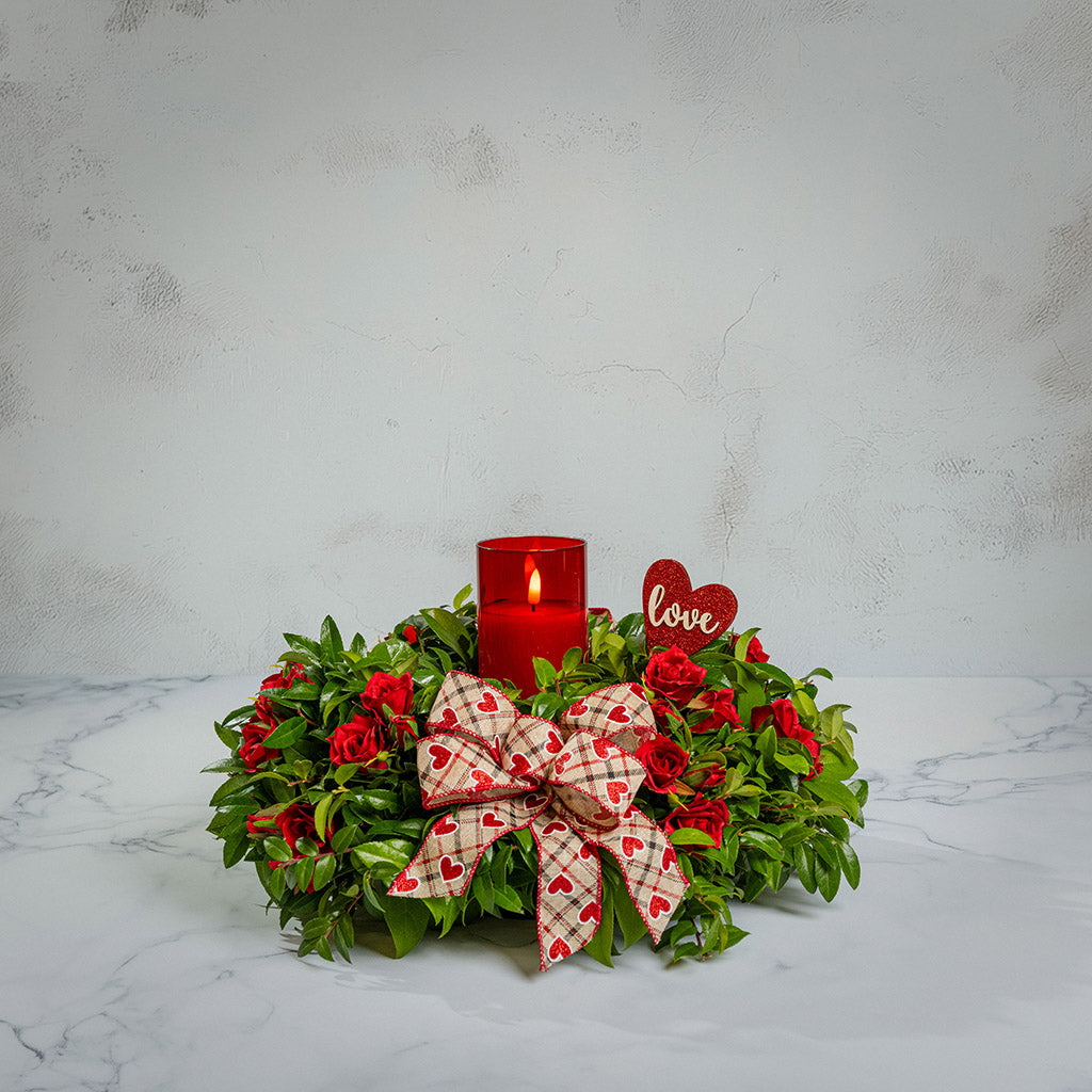 Fresh salal and green huckleberry with faux mini red roses, a red heart “love” sign, a red heart bow, and a red LED candle.