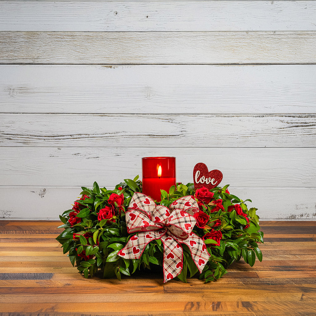 Fresh salal and green huckleberry with faux mini red roses, a red heart “love” sign, a red heart bow, and a red LED candle.