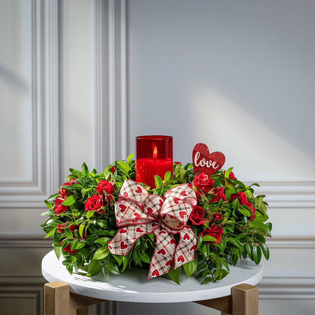 Fresh salal and green huckleberry with faux mini red roses, a red heart “love” sign, a red heart bow, and a red LED candle.