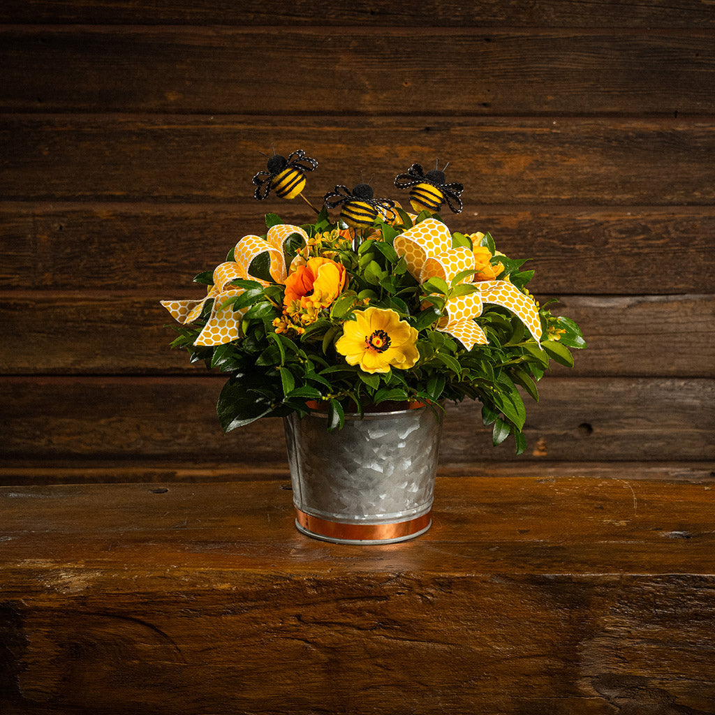 Fresh salal and green huckleberry with faux orange and yellow poppy flowers, faux yellow wax flowers, bumblebees, and a yellow honeycomb bow in a galvanized metal pot