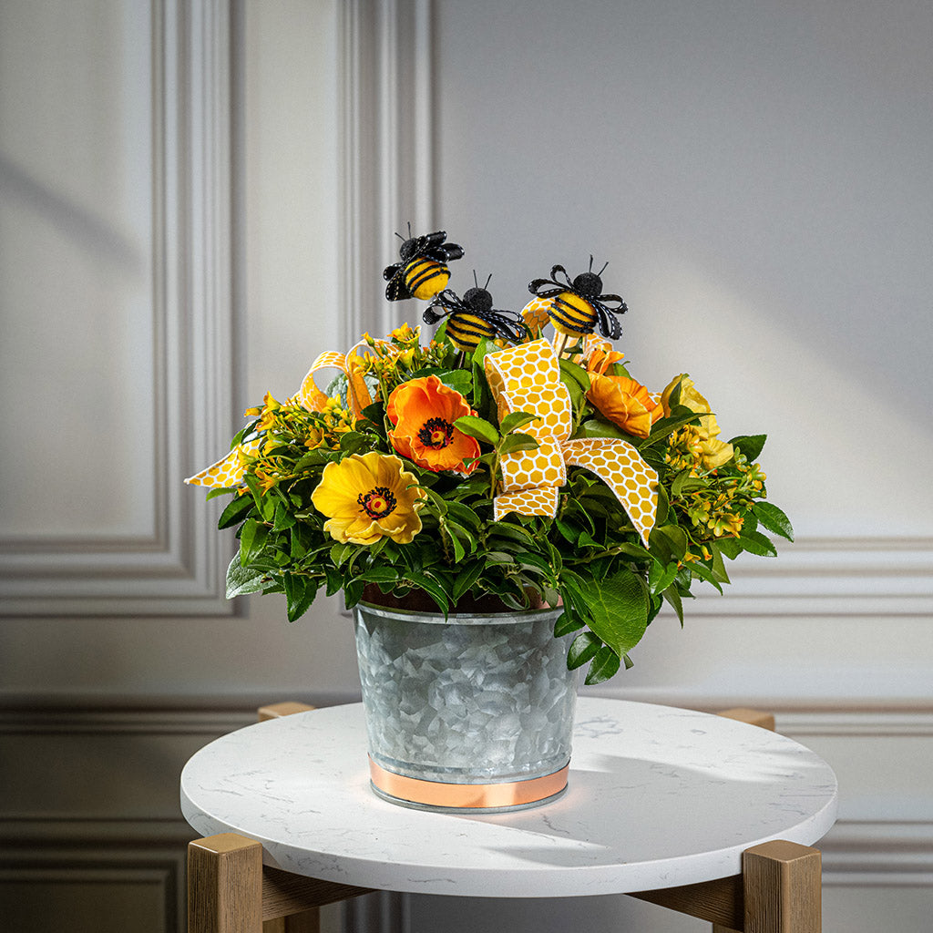 Fresh salal and green huckleberry with faux orange and yellow poppy flowers, faux yellow wax flowers, bumblebees, and a yellow honeycomb bow in a galvanized metal pot