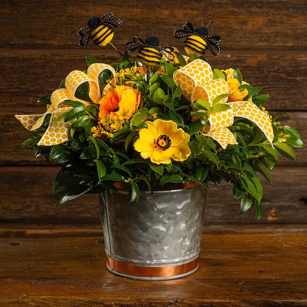 Fresh salal and green huckleberry with faux orange and yellow poppy flowers, faux yellow wax flowers, bumblebees, and a yellow honeycomb bow in a galvanized metal pot