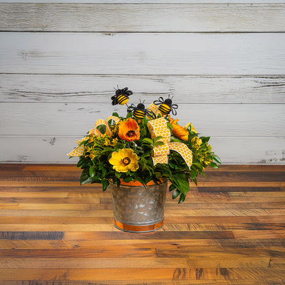 Fresh salal and green huckleberry with faux orange and yellow poppy flowers, faux yellow wax flowers, bumblebees, and a yellow honeycomb bow in a galvanized metal pot