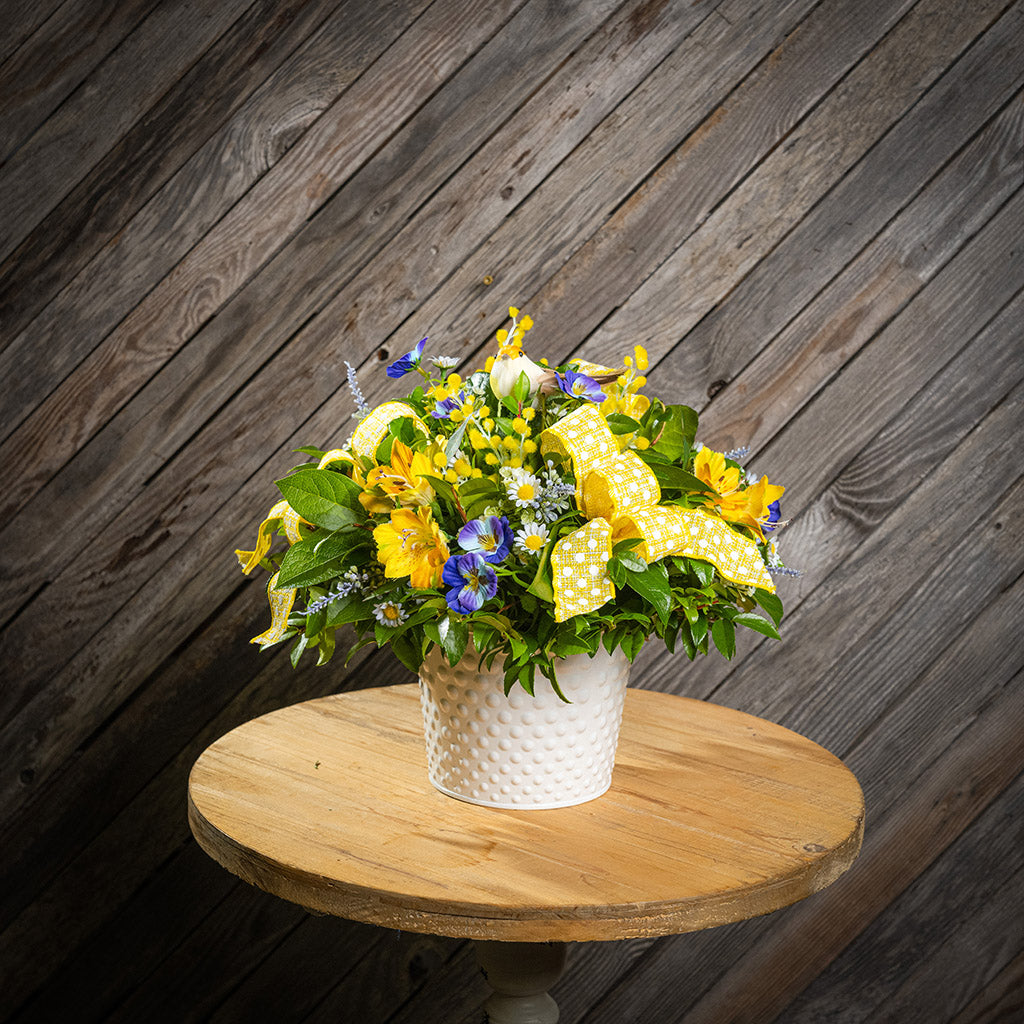 Fresh salal and green huckleberry with faux pansies, faux lavender, and faux yellow Alstromeria lilies, a yellow bird, and yellow polka-dot bows in a white metal pot