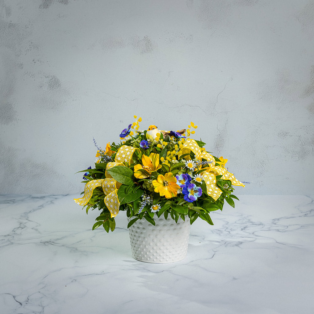 Fresh salal and green huckleberry with faux pansies, faux lavender, and faux yellow Alstromeria lilies, a yellow bird, and yellow polka-dot bows in a white metal pot