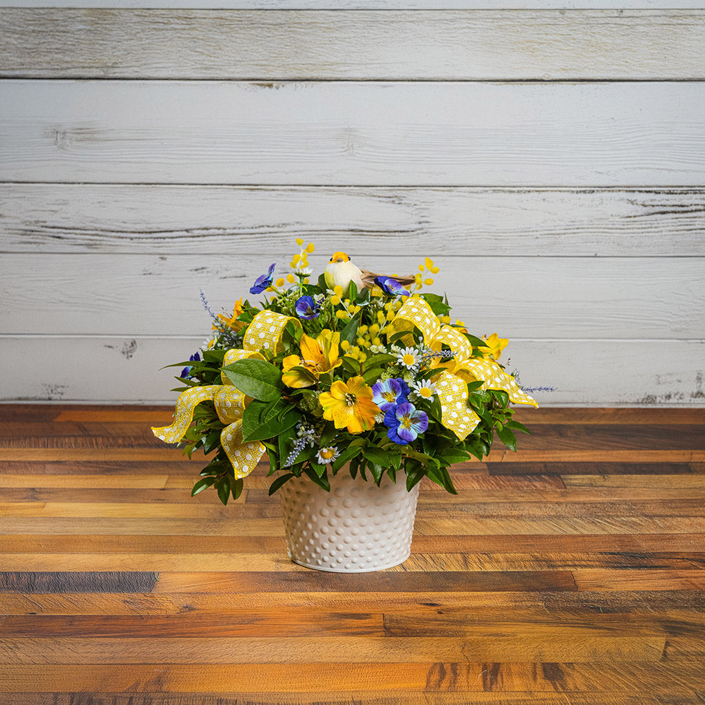 Fresh salal and green huckleberry with faux pansies, faux lavender, and faux yellow Alstromeria lilies, a yellow bird, and yellow polka-dot bows in a white metal pot