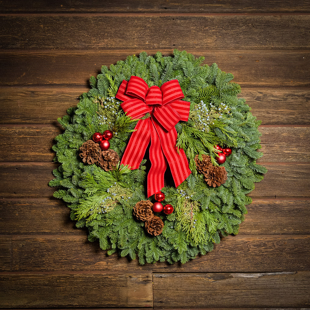 An evergreen wreath of noble fir, cedar, and juniper with pinecones, red ball ornaments, and a red-striped bow