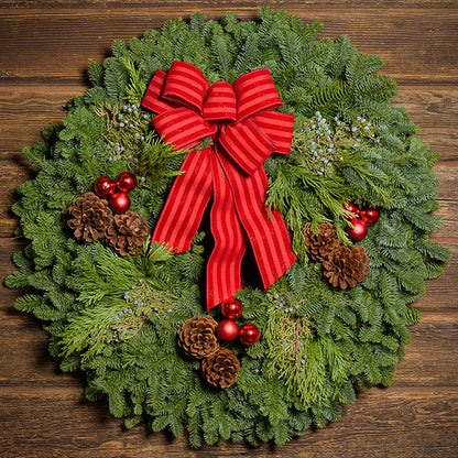 An evergreen wreath of noble fir, cedar, and juniper with pinecones, red ball ornaments, and a red-striped bow