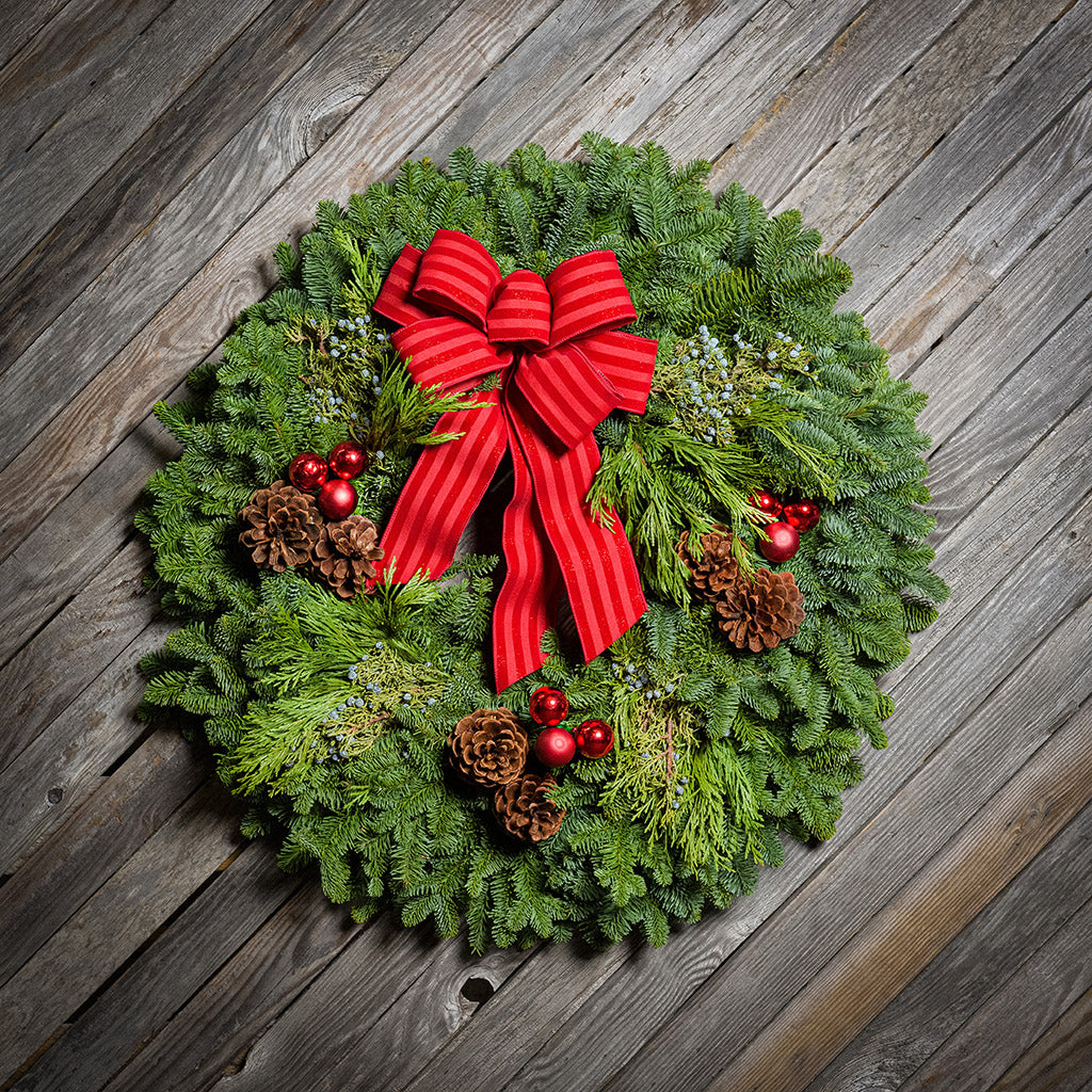 An evergreen wreath of noble fir, cedar, and juniper with pinecones, red ball ornaments, and a red-striped bow