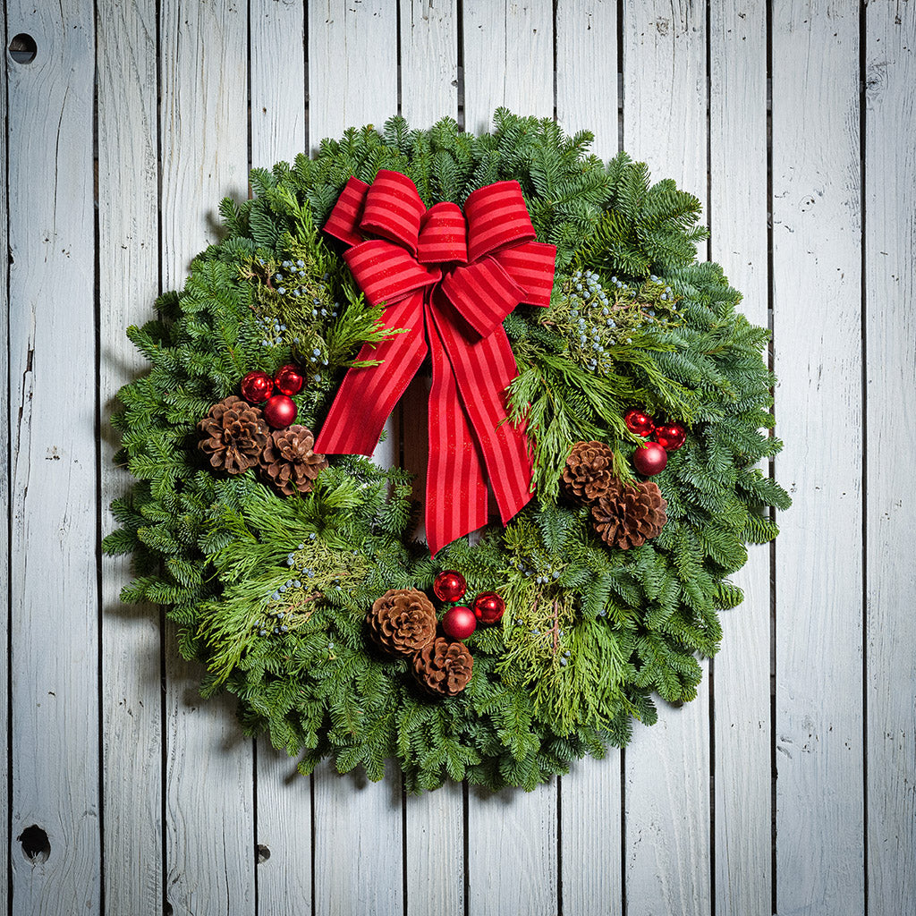 An evergreen wreath of noble fir, cedar, and juniper with pinecones, red ball ornaments, and a red-striped bow