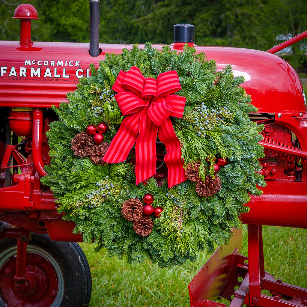 An evergreen wreath of noble fir, cedar, and juniper with pinecones, red ball ornaments, and a red-striped bow