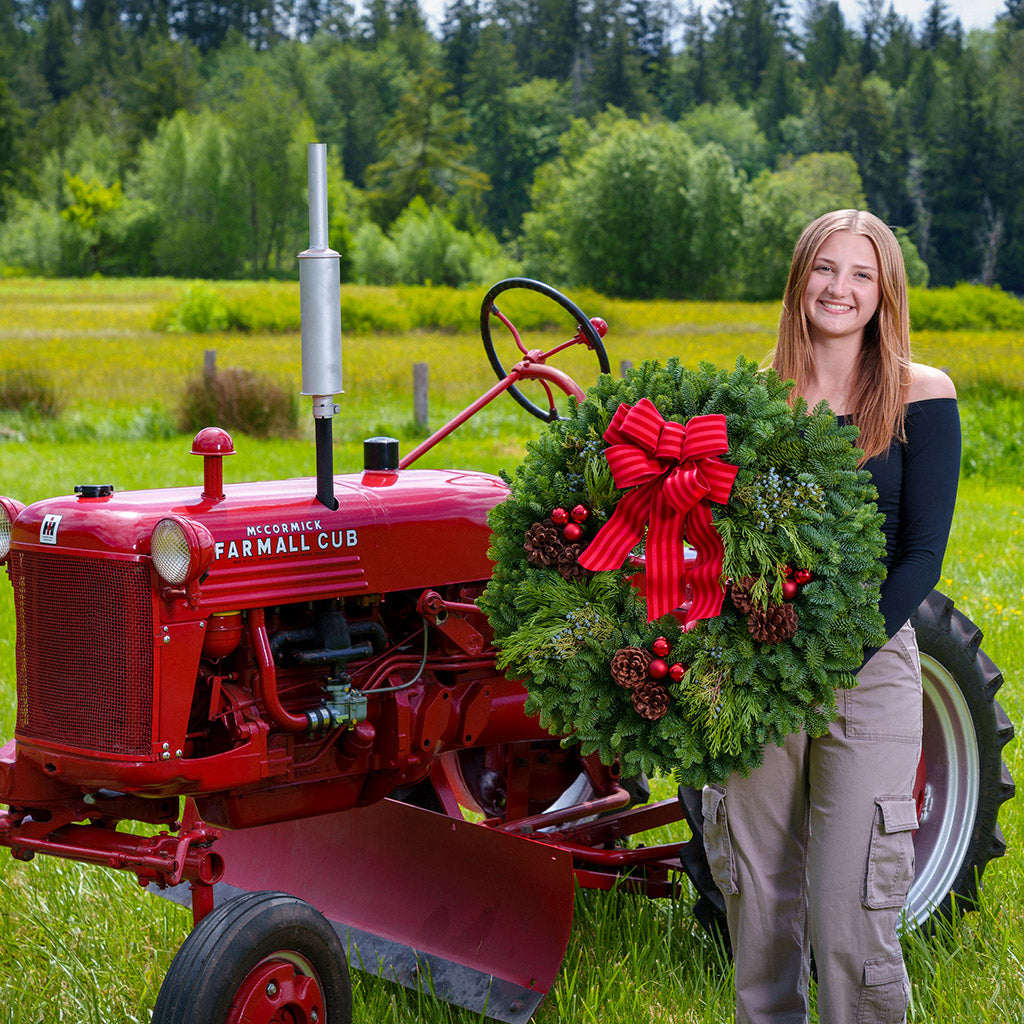 An evergreen wreath of noble fir, cedar, and juniper with pinecones, red ball ornaments, and a red-striped bow