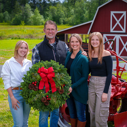 An evergreen wreath of noble fir, cedar, and juniper with pinecones, red ball ornaments, and a red-striped bow