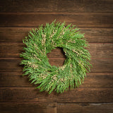 A dried wreath made of natural green oats, preserved fern, and ivory flax pods hung on a dark wood background.