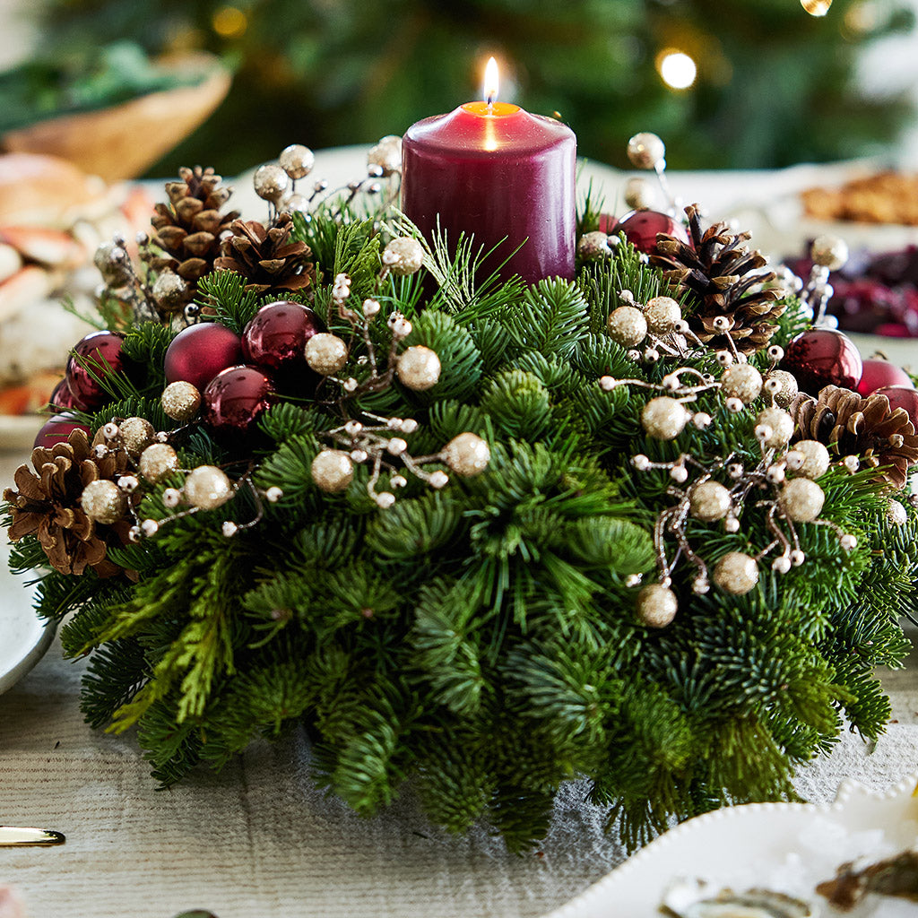 Centerpiece made of verdant fresh noble fir, white pine, and incense cedar decorated with glittery beads and shiny burgundy balls, natural pinecones and a deep burgundy pillar candle