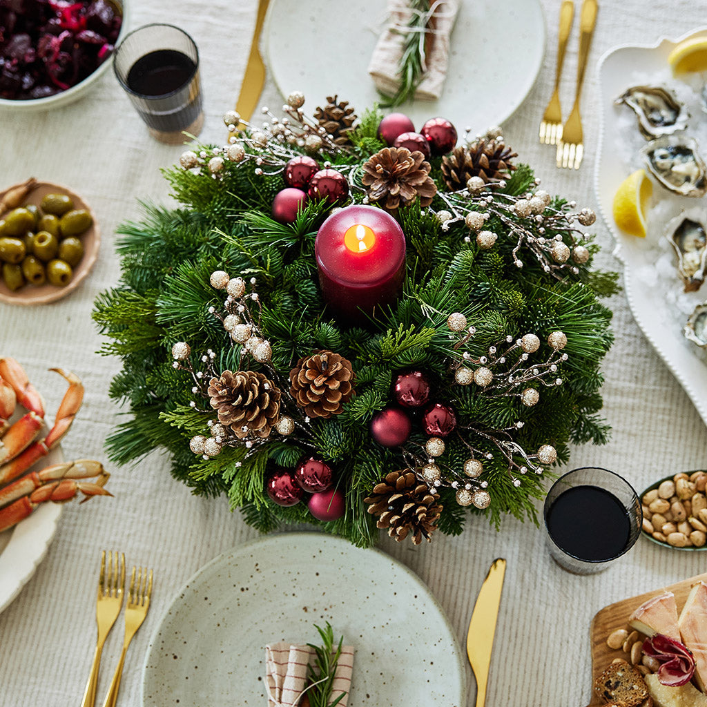 Centerpiece made of verdant fresh noble fir, white pine, and incense cedar decorated with glittery beads and shiny burgundy balls, natural pinecones and a deep burgundy pillar candle