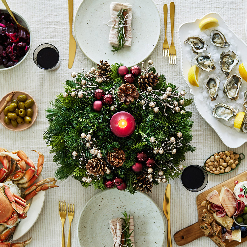 Centerpiece made of verdant fresh noble fir, white pine, and incense cedar decorated with glittery beads and shiny burgundy balls, natural pinecones and a deep burgundy pillar candle