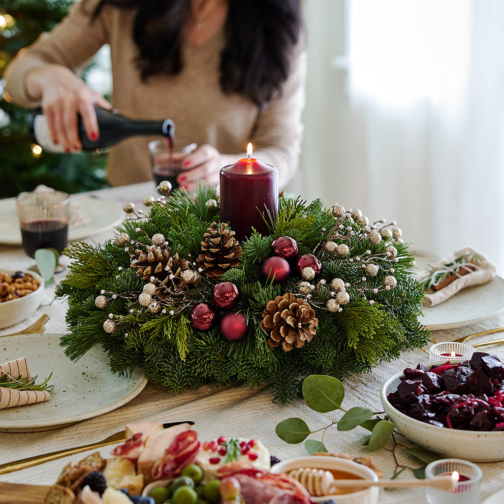 Centerpiece made of verdant fresh noble fir, white pine, and incense cedar decorated with glittery beads and shiny burgundy balls, natural pinecones and a deep burgundy pillar candle