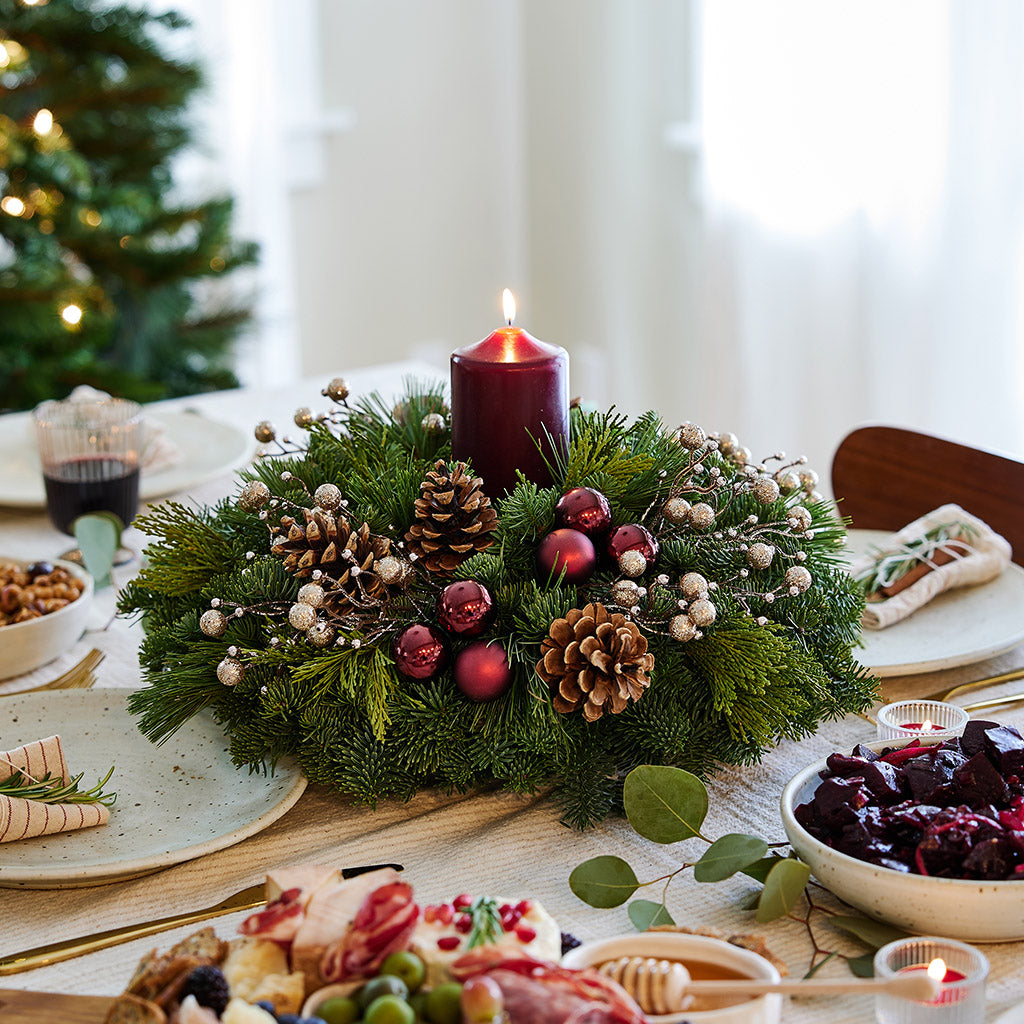 Centerpiece made of verdant fresh noble fir, white pine, and incense cedar decorated with glittery beads and shiny burgundy balls, natural pinecones and a deep burgundy pillar candle