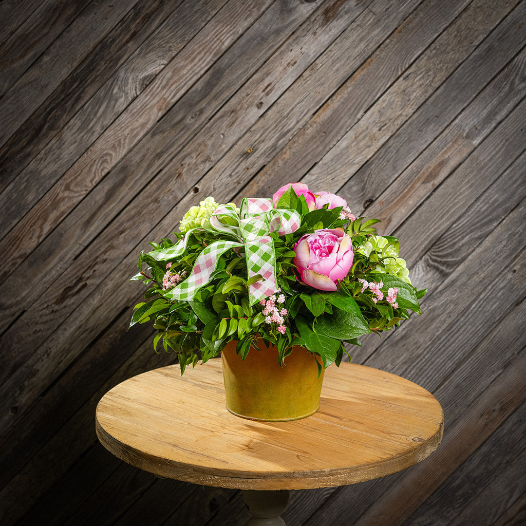 Fresh salal and green huckleberry with faux green hydrangeas, faux pink peonies, faux pink baby’s breath flowers, and plaid mauve and a sage-green bow in a spring-colored metal pot