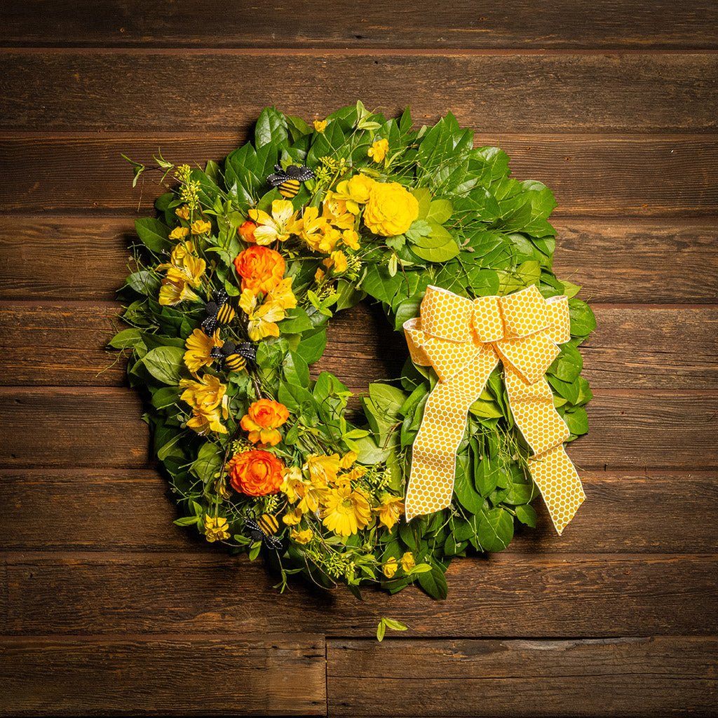 Fresh salal and sweet huckleberry with faux spring wildflowers, faux gerbera daisies, faux yellow and orange ranunculus flowers, honey bees, and a yellow honeycomb-patterned bow