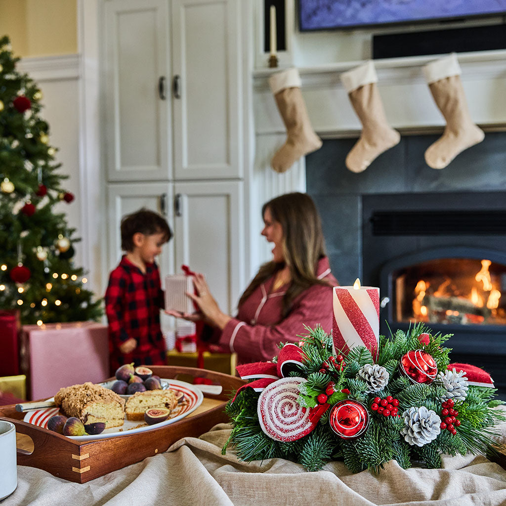 A cozy holiday living room scene with a woman and a young boy by a fireplace, one holding a wrapped gift. In the foreground, a festive holiday wreath with red-and-white striped candles, pinecones, and red berries sits on a table beside a tray of baked goods.