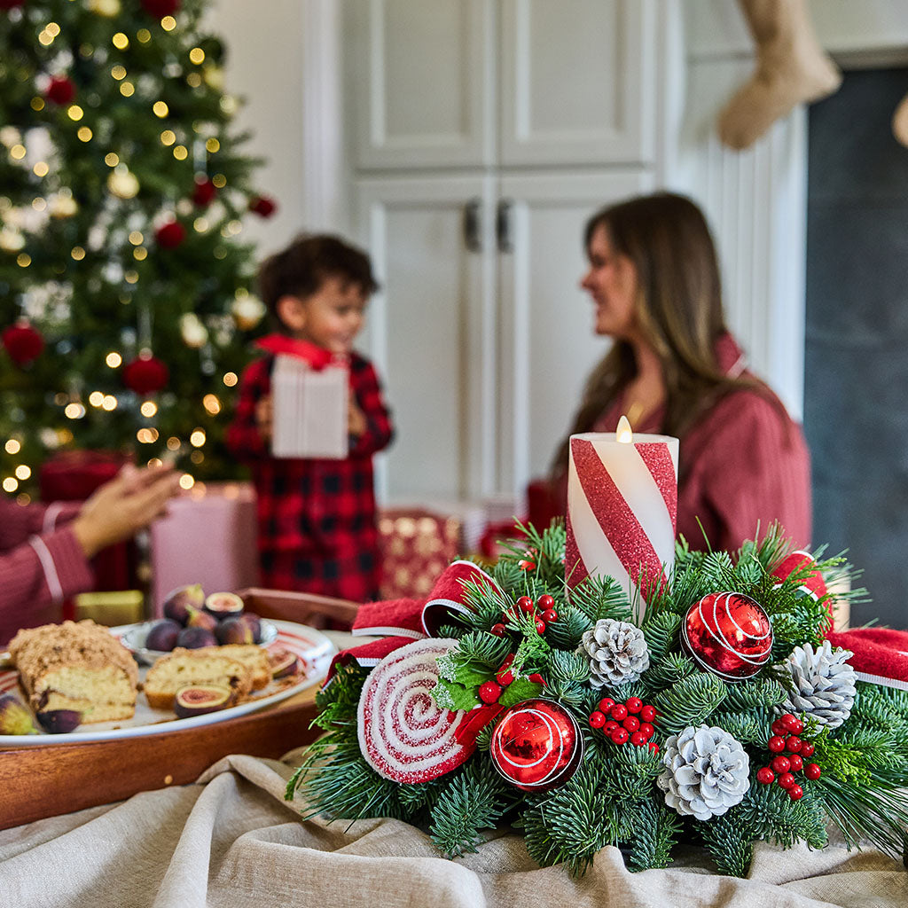 A festive Christmas holiday table centerpiece featuring a green pine garland with red ornaments, silver pinecones, red berries, and a red-and-white striped candle, set on a beige tablecloth in a cozy living room with a blurred Christmas tree and family in the background.