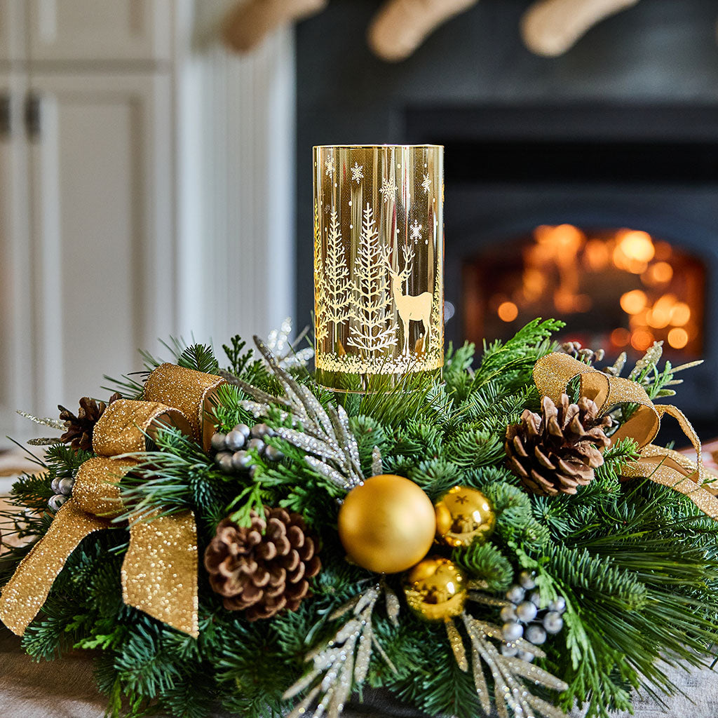A festive gold holiday candle holder with a deer and tree design, nestled in a green pine wreath with gold ribbons, pinecones, and ornaments in front of a warm fireplace.