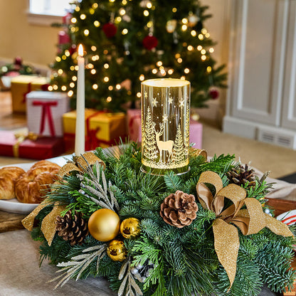 A festive Christmas centerpiece with a glowing gold cylindrical lantern surrounded by green pine branches, gold ribbons, pinecones, and gold ornaments, set on a table in a decorated living room with a Christmas tree and wrapped gifts in the background.