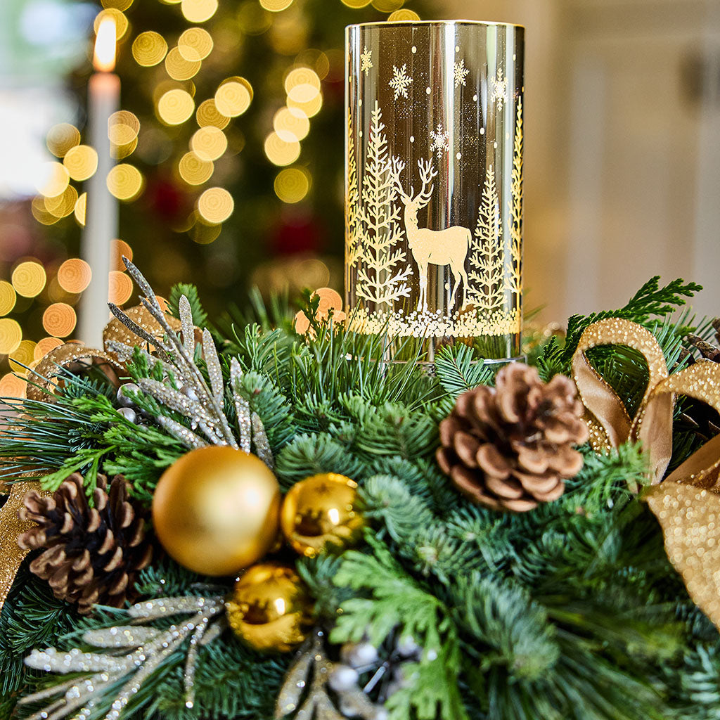 A cylindrical glass candle holder with a gold deer and tree design, displayed on a green Christmas wreath with pinecones, gold ornaments, and a blurred festive background.