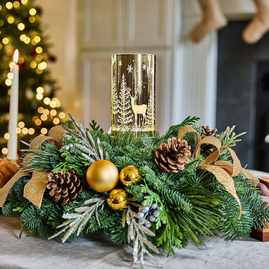 A festive Christmas centerpiece with a gold cylindrical candle holder featuring a deer and tree design, surrounded by green pine branches, gold ornaments, pinecones, and a gold ribbon, set on a table in a softly blurred holiday interior.