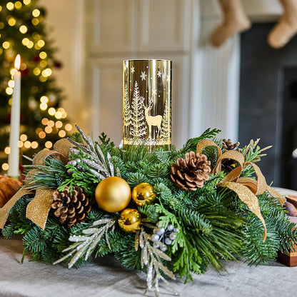 A festive Christmas centerpiece with a gold cylindrical candle holder featuring a deer and tree design, surrounded by green pine branches, gold ornaments, pinecones, and a gold ribbon, set on a table in a softly blurred holiday interior.